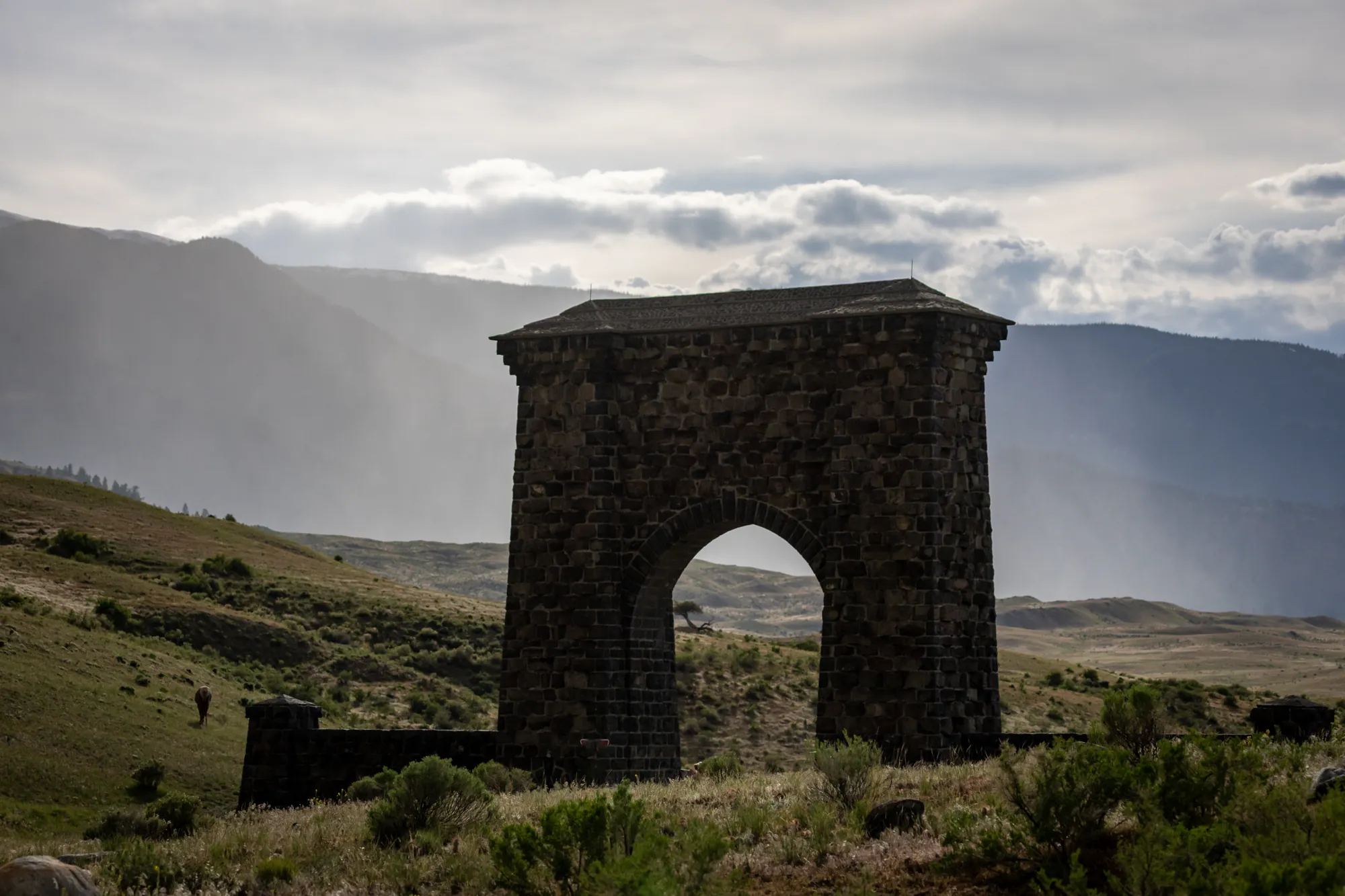 The Roosevelt Arch, a tall basalt stone gateway on the sagebrush plain at Yellowstone's North Entrance under a stormy sky
