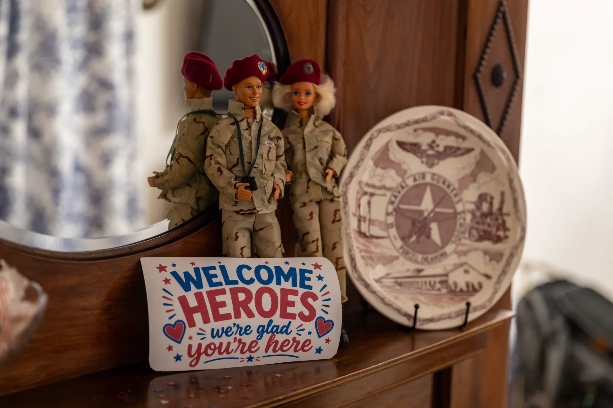 A "Welcome Heroes, we're glad you're here" placard on a shelf beside military figurines and a commemorative plate